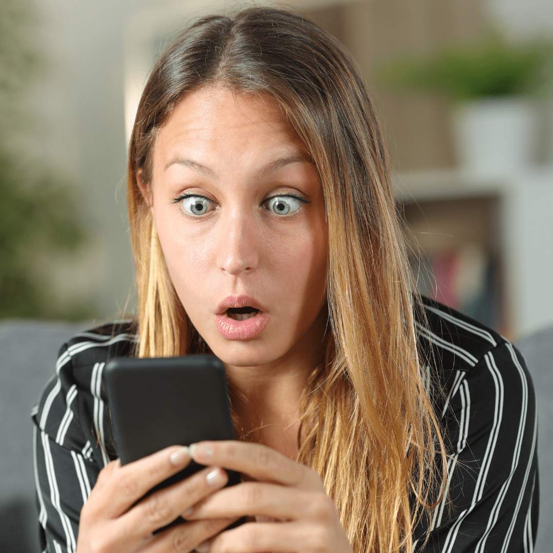 Shocked woman with wide eyes and open mouth staring at her smartphone in disbelief, seated indoors with a blurred background. indicated sticker shock at ride share hidden fees